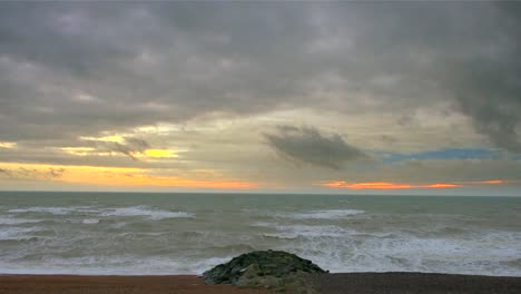 Der-Graue-Ozean-Trifft-Auf-Den-Kiesstrand-Unter-Einem-Schweren-Himmel,-Der-Von-Der-Untergehenden-Sonne-Orange-Gefärbt-Wird