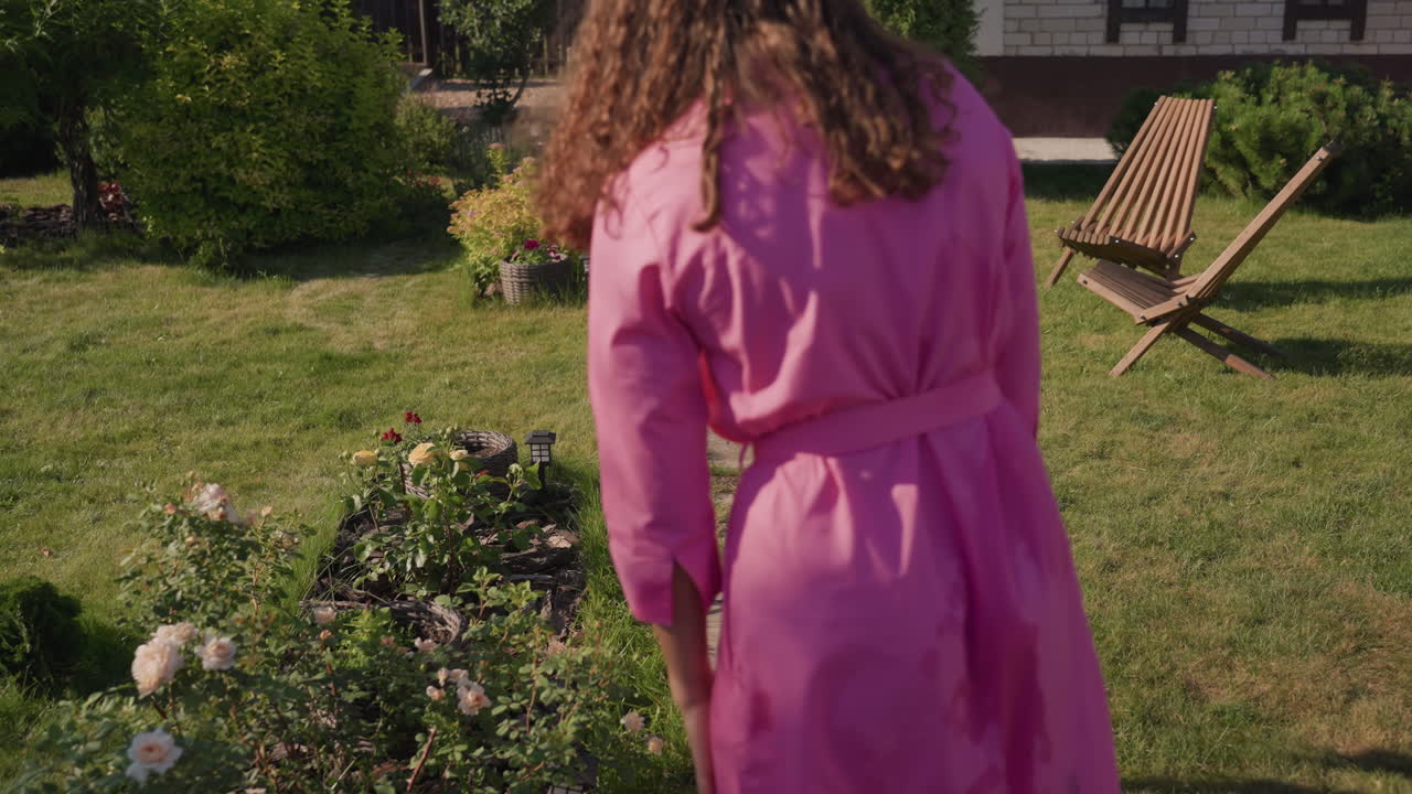 Youthful Flower Play, Girl Enjoys Spraying Garden Blooms Under Bright Sunny Sky While Adult Observes, Young Girl Mischievously Sprays Flowers In Sunny Garden As Adult Watches Nearby From Patio