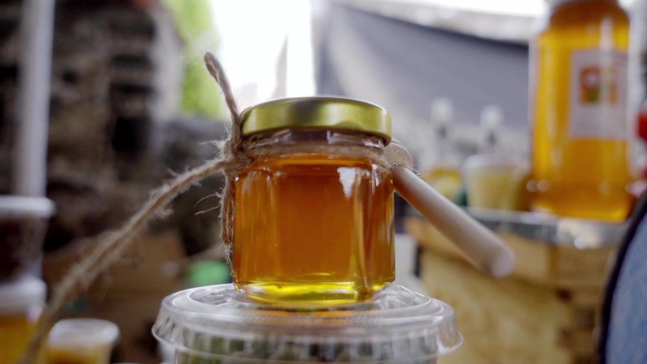 Vista ravvicinata di un piccolo barattolo di vetro pieno di miele dorato, legato con spago rustico e con un mestolino di legno appoggiato sopra, con luce calda che evidenzia la consistenza e la dolcezza naturale in un ambiente cucina