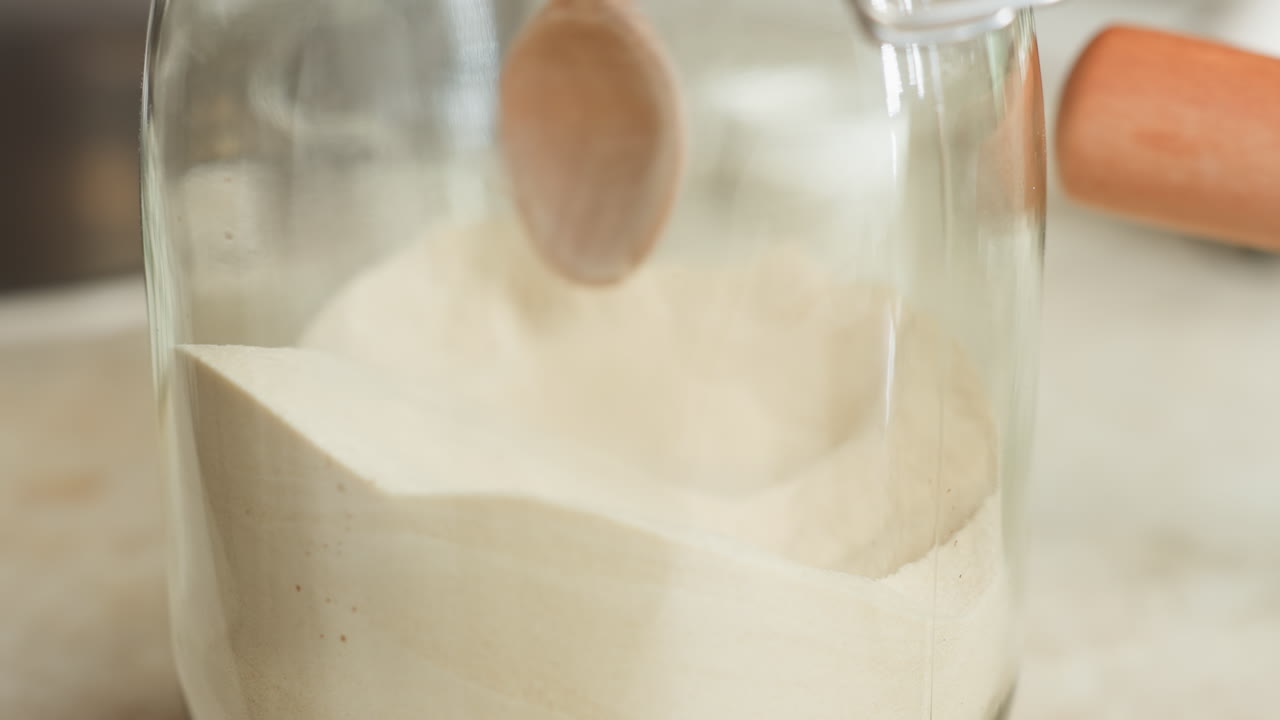Close up of corn flour being picked up with wooden spoon inside transparent glass container, showing texture of flour and kitchen surface in soft natural light during home cooking