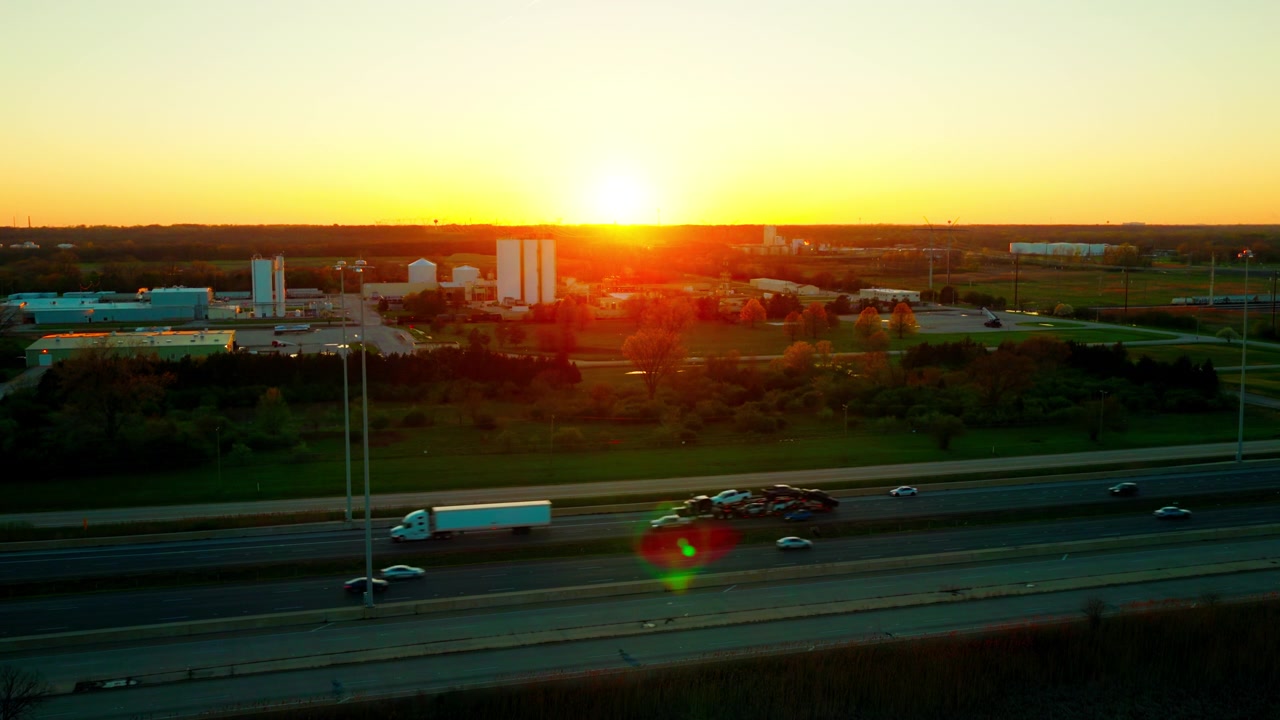 Semi truck car hauler on Illinois interstate at golden hour. Mixed traffic, light industrial campus, saffron sunset sky. Automotive logistics B-roll.