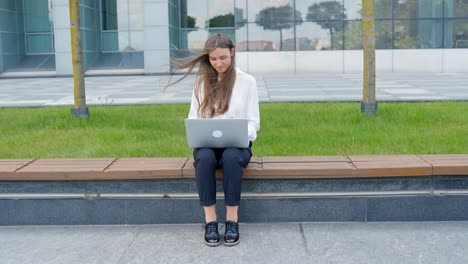 sonrisa morena atractiva mientras trabaja con una computadora portátil al aire libre cerca de la oficina