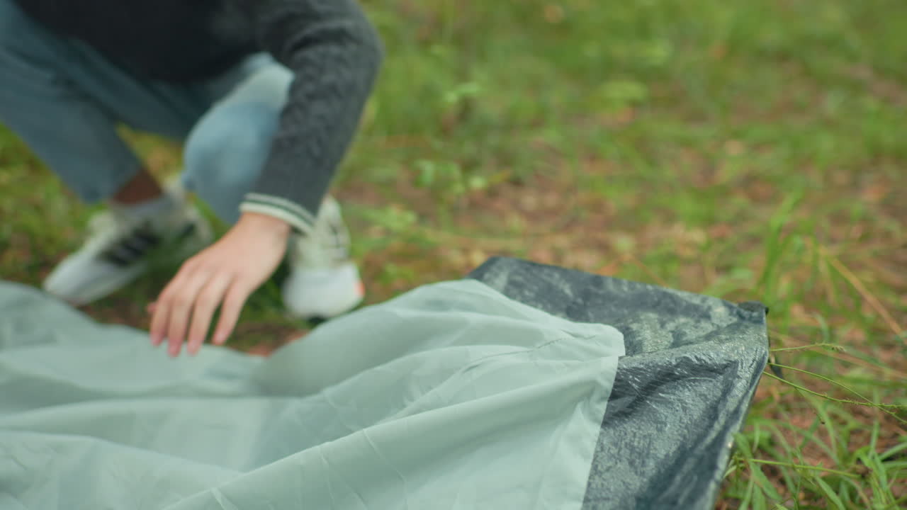 Man in green knitted sweater squatting on forest ground with warm smile directed at someone in background while preparing camping tent in natural woodland