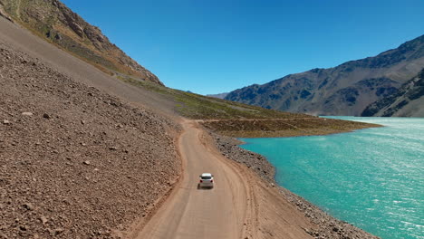 white car driving along scenic dirt road beside turquoise embalse el yeso reservoir in andes mountains, travel and adventure in cajón del maipo, chile. aerial tracking