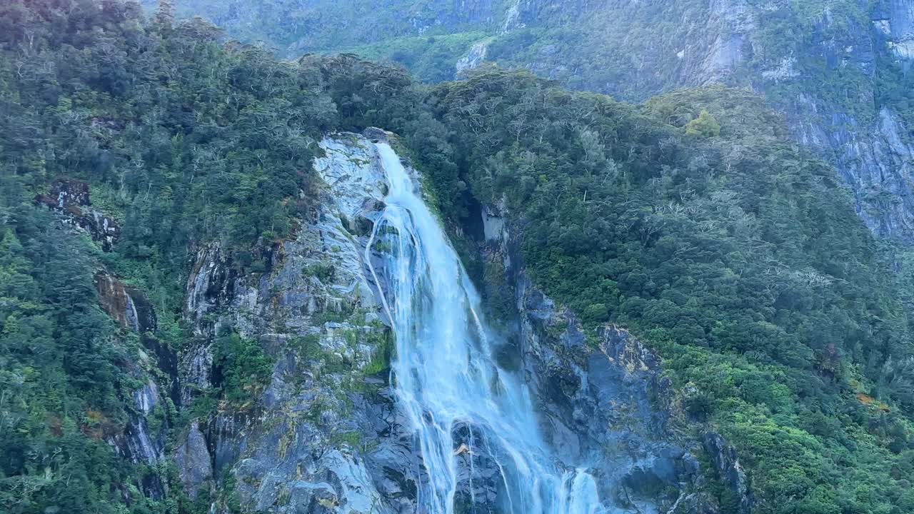 Plavba kolem velkého vodopádu v Milford Sound, Národní park Fiordland, Nový Zéland. Tyčící se útesy, kaskádovitá voda a mlhavé okolí vytvářejí dramatický a malebný výhled na fjord.