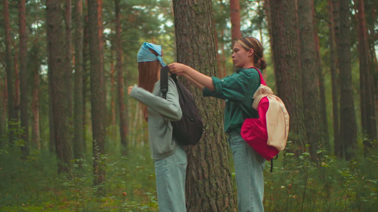 joven con mochila roja ayuda a su amiga a ajustar su bolsa negra en un entorno pacífico del bosque, la amiga con bufanda azul se da la vuelta y sonríe cálidamente antes de caminar juntos