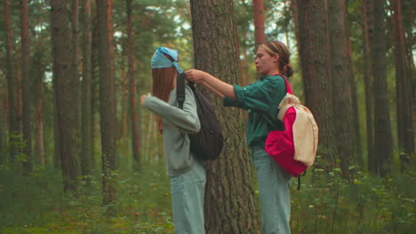 young woman in red backpack helps her friend adjust her black bag in a peaceful forest setting, friend in blue scarf turns and smiles warmly before walking together