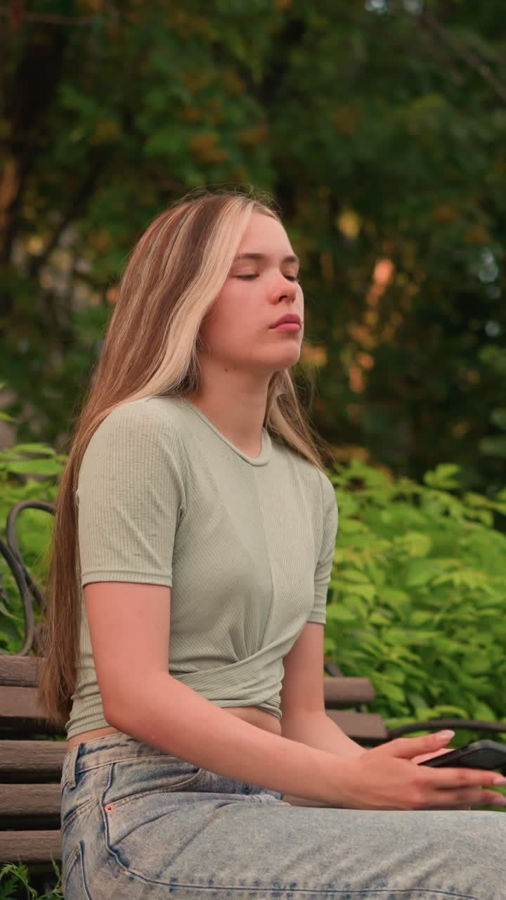 mujer joven al aire libre, sosteniendo el teléfono en su mano derecha y mirando lejos pensativo y se sienta en el banco de madera, con un traje casual con crop top y vaqueros, rodeado de exuberante vegetación