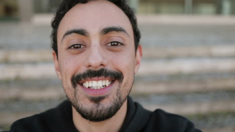 Close-up-shot-of-young-Arabic-mans-face-with-dark-curly-hair-and-beard-in-black-hoodie-sitting-on-stairs-outside