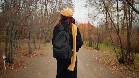 back view of lady with red curly hair walking through forest path surrounded by autumn foliage, carrying black bag and wearing yellow beret