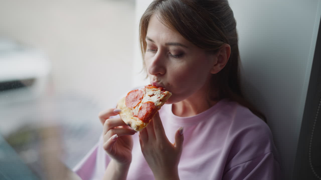 Woman wearing purple shirt sitting by window holding toasted bread looking thoughtfully at it before eating, natural light from outside creating calm reflective atmosphere, background
