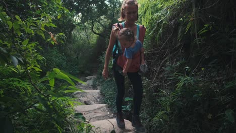 family hiking. mother hikes with baby in the tropical forest. young woman hiker carries her baby in wrap sling and walks in the forest