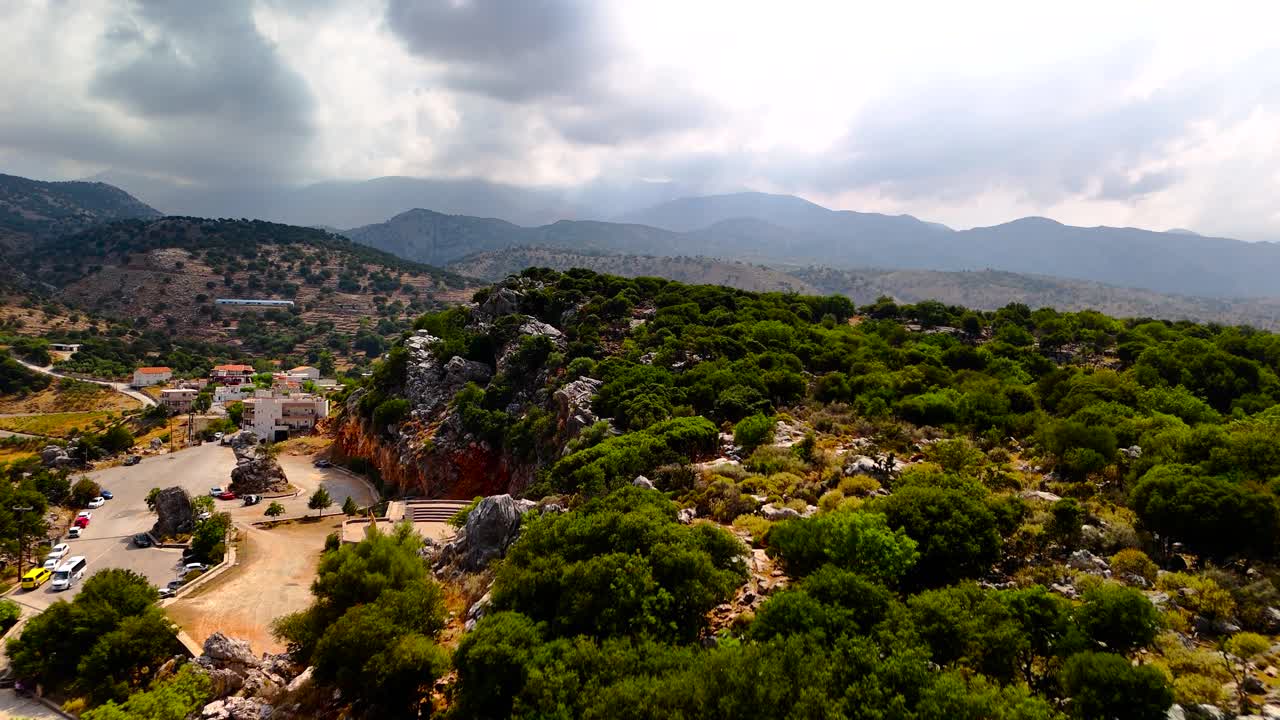 A beautiful landscape shows rolling hills, scattered trees, and a small village nestled among nature. The sky is overcast, giving a serene atmosphere to the rural setting