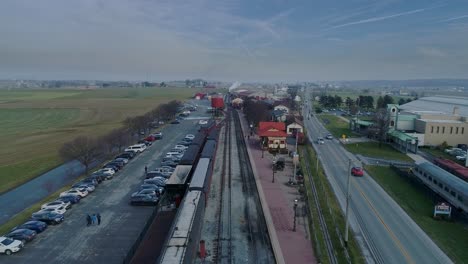 An-Aerial-View-of-a-Train-Station,-with-a-Steam-Passenger-Train,-Pulling-into-the-Station,-Blowing-Smoke,-in-Slow-Motion-on-a-Partially-Sunny-Day