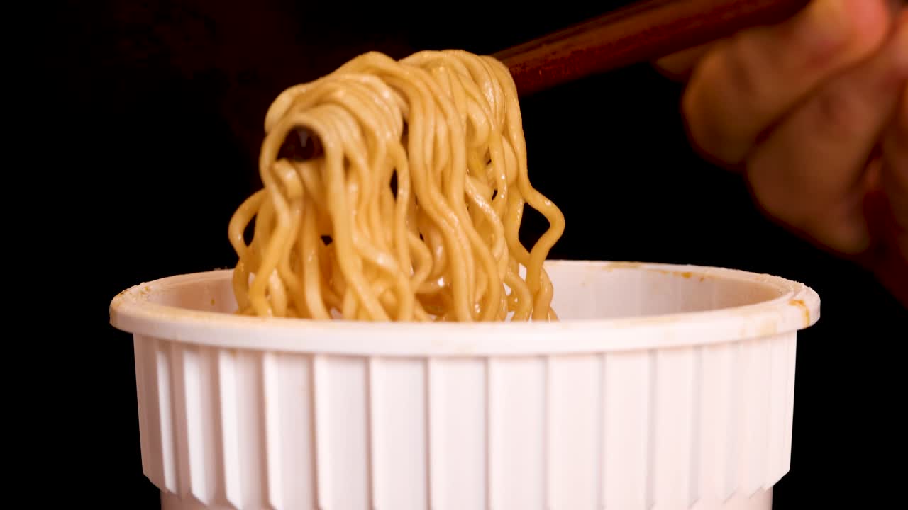 A hand skillfully lifts cooked instant noodles from a white cup using wooden chopsticks, set against a black background with warm, even lighting and close-up framing