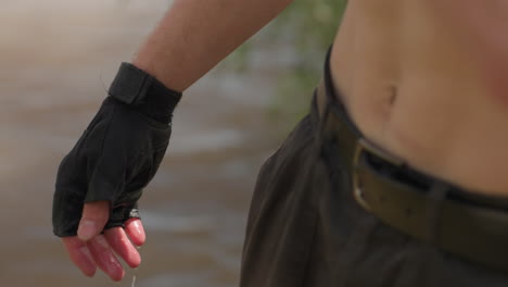 close view of male adventurer bending near river washing face, water droplets falling from chin, white headband over brown hair, black tattoo visible on neck, natural light highlighting quiet moment