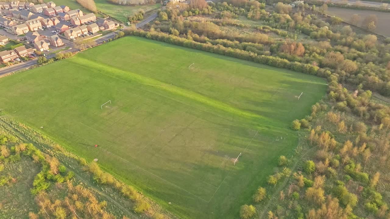 Expansive green football pitch bordered by dense trees and a nearby residential area in warm evening light in Wombwell, South Yorkshire, England, static drone shot