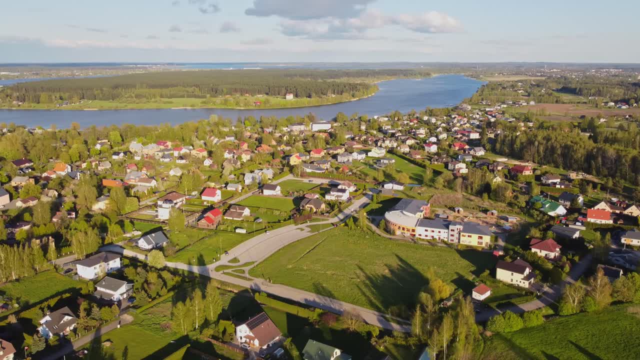 Aerial view revealing verdant Katlakalns landscape, colorful housing nestled among spring greenery, Lielupe river winding through Latvian countryside under bright blue sky
