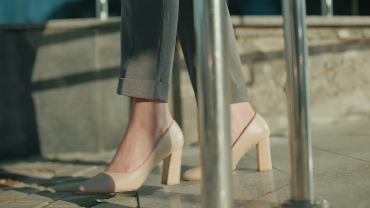Leg view of young woman walking on interlocked pavement wearing beige heels and grey trousers, sunlight casting shadows along sidewalk with stone wall and urban railings in background