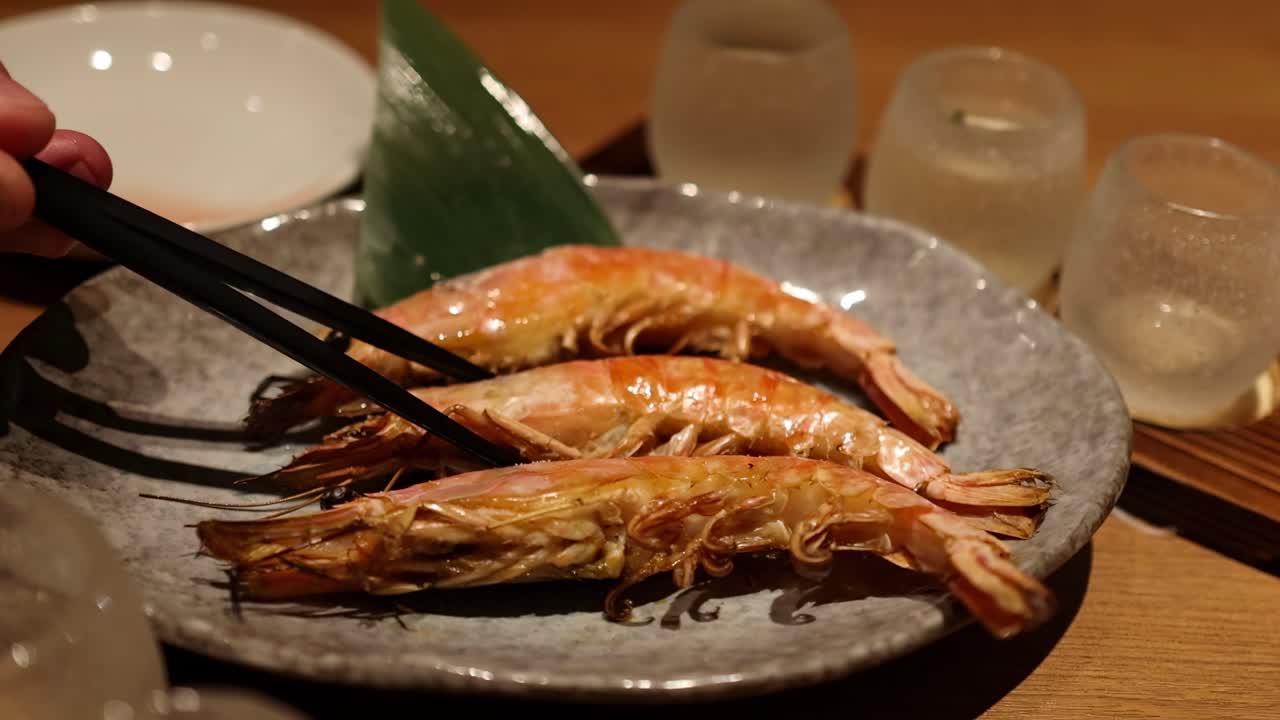 Close-up of chopsticks picking up grilled shrimp from a stone plate with decorative leaf.