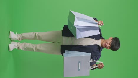 vista completa de un hombre de compras asiático con bolsas de compras sonríe a la cámara mientras está de pie frente a la pantalla verde