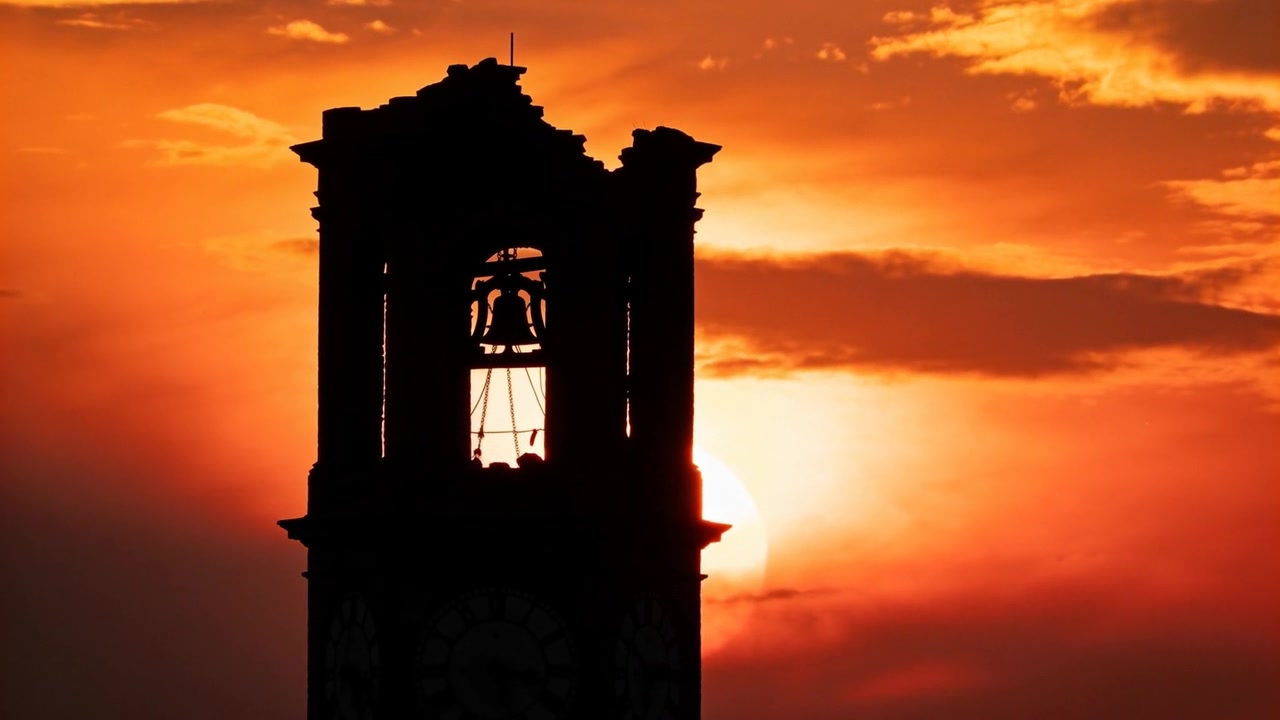 Sole che scivola dietro il campanile al tramonto, rivelando campana in ghisa, batacchio e corde.