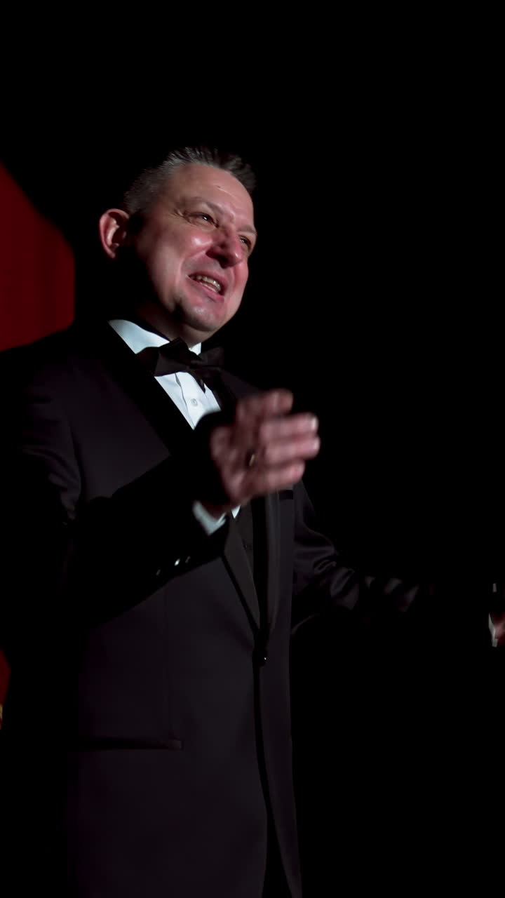 Male host talking on stage. Performer man in black suit standing on dark stage at spotlight and speaking with gestures in the theater. Circling camera. Vertical video
