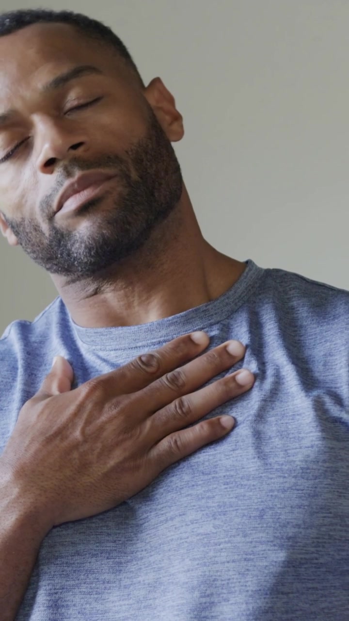 Video verticale: Uomo che respira si porta la mano destra al petto in studio, indossa una maglietta blu mélange.