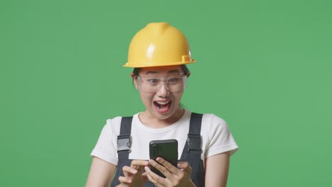 close up of asian woman worker wearing goggles and safety helmet looking at smartphone, smiling, and saying wow while standing in the green screen background studio