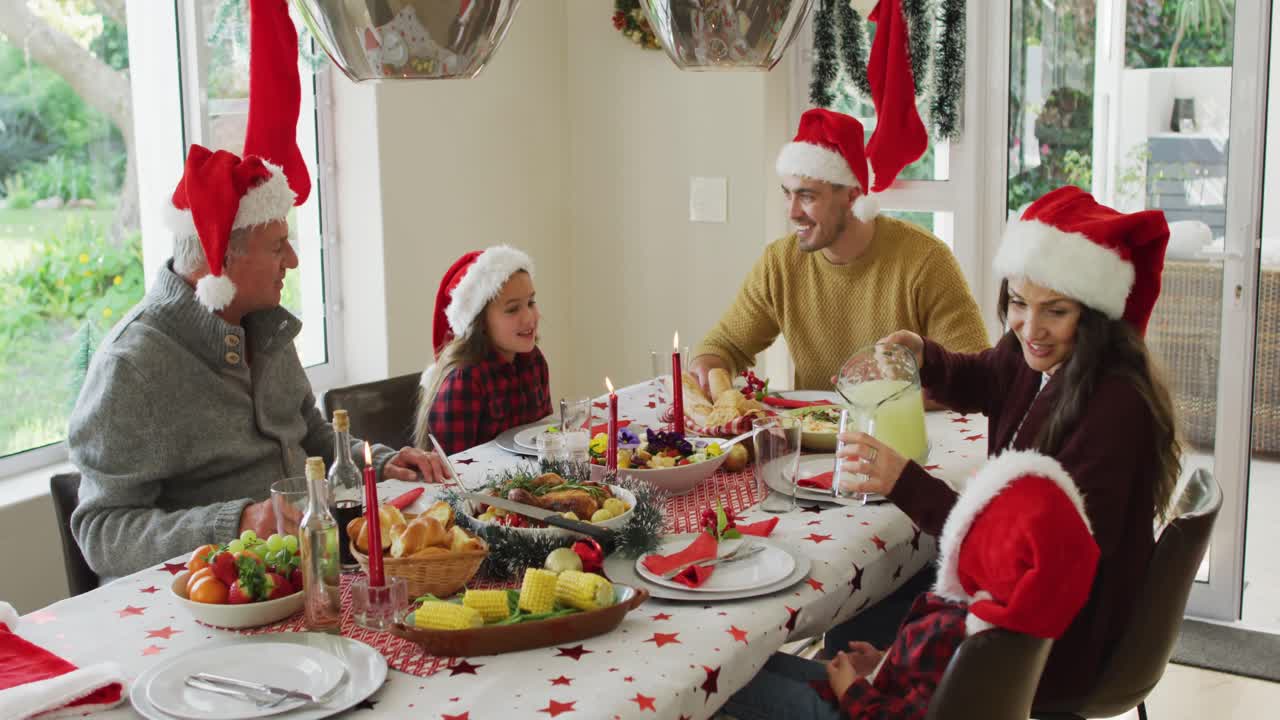 familia caucásica feliz de varias generaciones con sombreros de papá noel, teniendo la comida de navidad