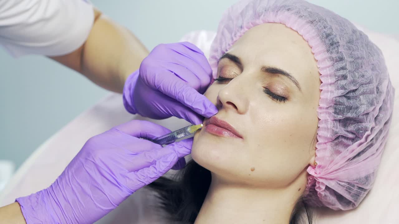 Beautiful woman is getting an injection in her lips in clinic. Cosmetologist in gloves pricks female's lips with syringe