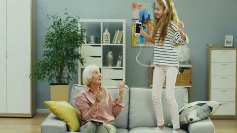 joyful teenager girl in headphones listening to the music and jumping on the couch in the living room while her good looking grandmother sitting beside her, trying to talk to her and stop. inside.