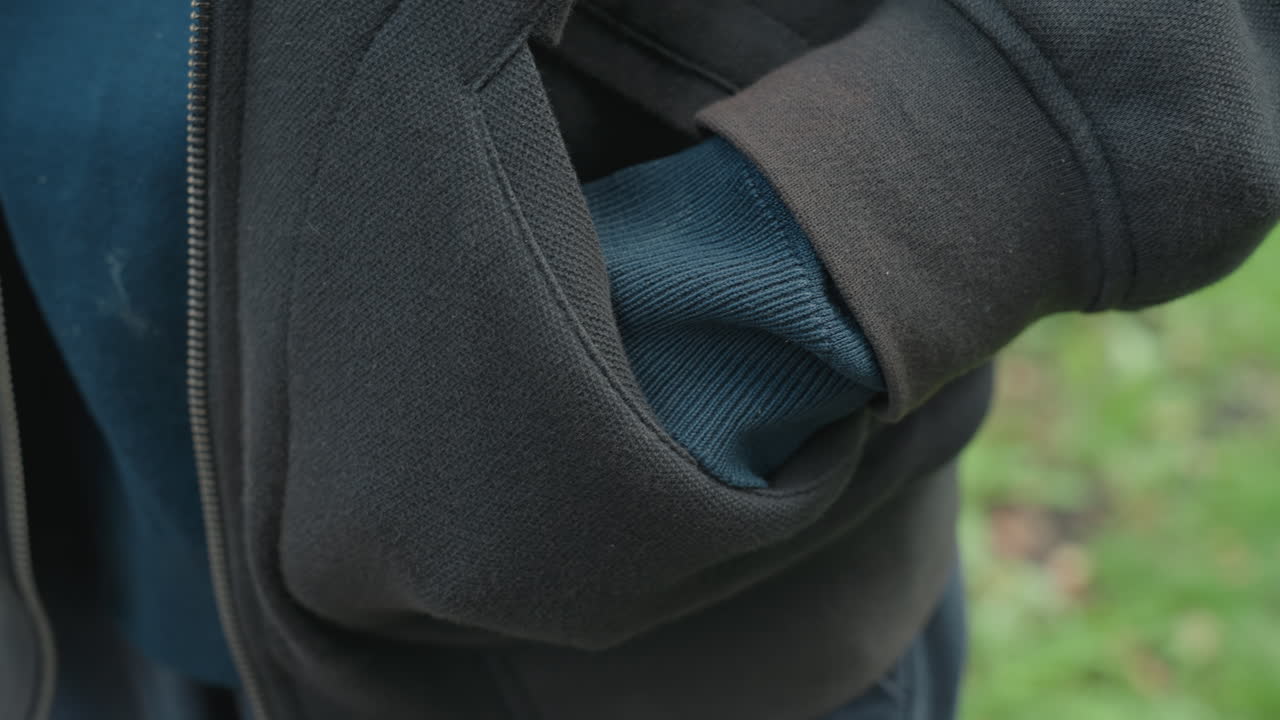 Punho cerrado e mão escondida perto do bolso, close-up do punho de malha e do bolso de fleece gasto, energia nervosa sutil de um transeunte capturada em pequenos gestos, dia nublado na trilha do parque, textura e atmosfera destacadas