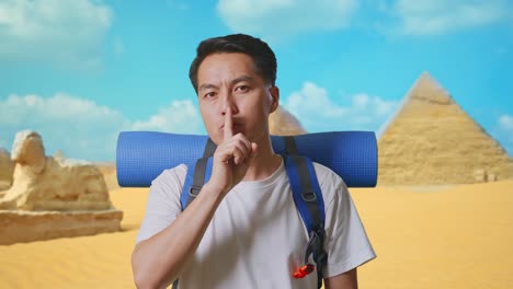 close up of asian male hiker with mountaineering backpack looking at camera and making shh gesture while traveling in pyramid of giza