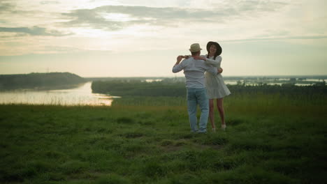 a man spins his wife around on a grassy hill beside a peaceful lake at sunset. the man, dressed in a white shirt, hat, and jeans, shares a tender moment with his wife, as she embraces him