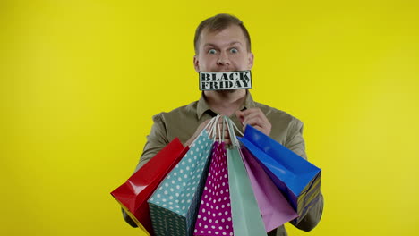 surprised man showing shopping bags and black friday inscription in his mouth. yellow background