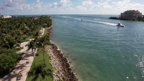 Drohnenclip-Des-Yachthafens-Von-Miami,-Der-Einen-Von-Palmen-Gesäumten-Gehweg-Und-Blaues-Flusswasser-Mit-Booten-Zeigt,-Die-Sich-Durch-Den-Rahmen-Bewegen,-Und-Das-Offene-Meer-Im-Hintergrund