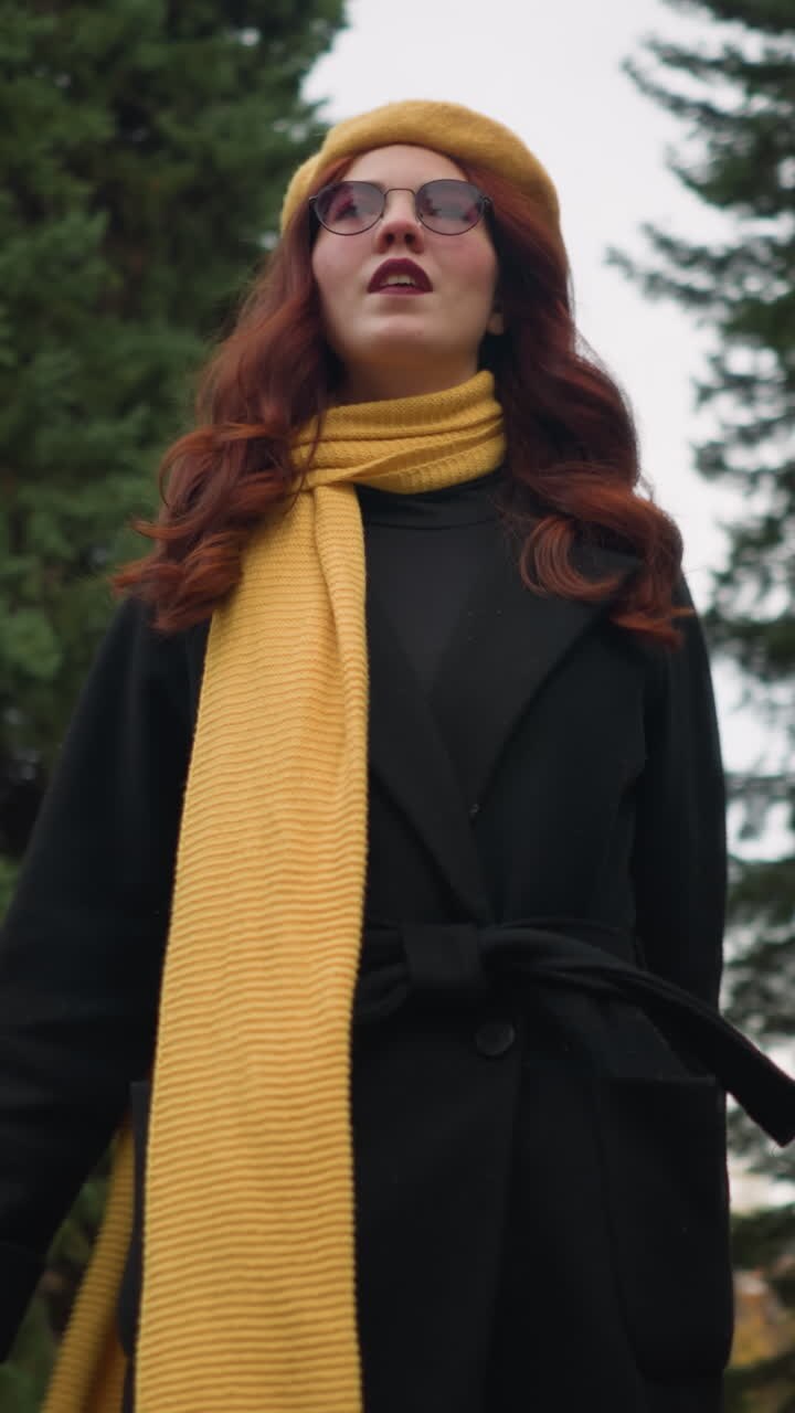 Una signora con i capelli rossi e ricci si gira gioiosamente, con una mano sulla testa, indossando un basco giallo e una sciarpa, ondeggiando mentre si gode la vivace atmosfera del giardino.