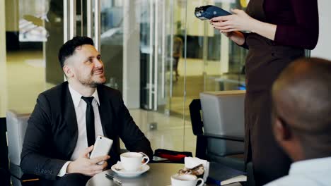 smiling bearded caucasian businessman sitting with his african american partner and paying online bill using his smartphone in cafe. cheery waitress using cash terminal.