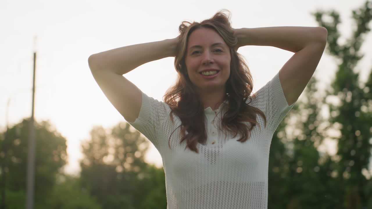 portrait female parent on picnic outing smiling with hands behind head, sun backlight, green trees blurred background, relaxed joyful mood, warm light creating glow around hair and face