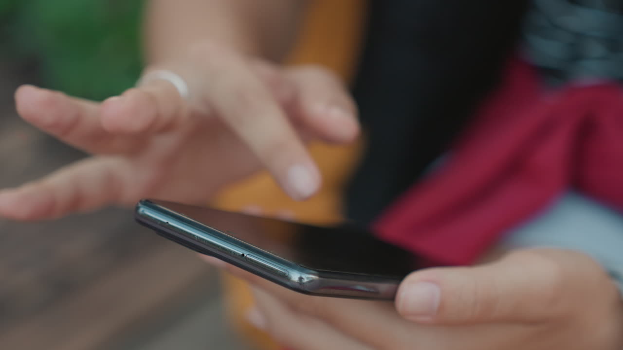 Primo piano di mani femminili che scorrono il telefono all'aperto, giacca colorata e anello visibili, scorrimenti e tocchi rapidi mentre controllano app e notifiche in un ambiente casual da parco con un'atmosfera energica.