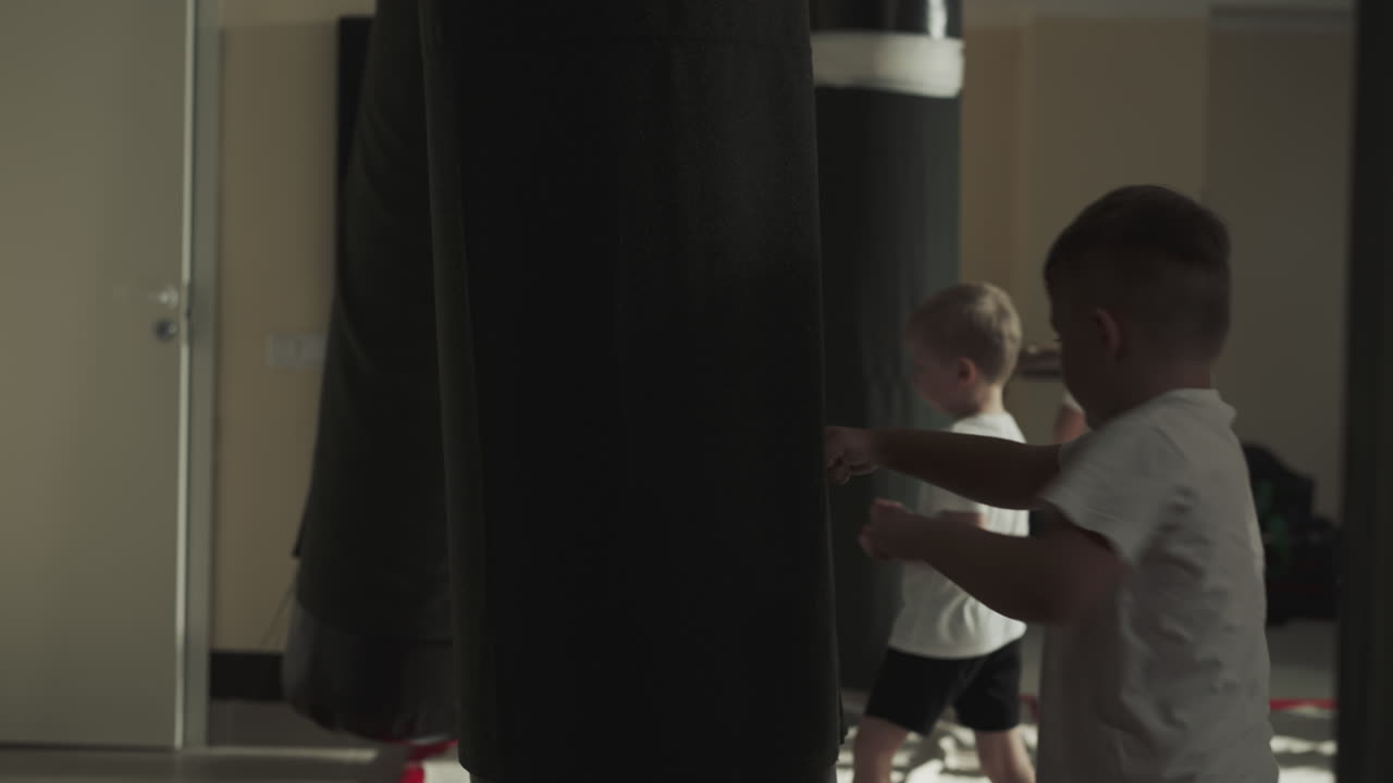 Child punches large hanging bag in sports club. Toddler boy hits at exercising heavy equipment in sports school slow motion. Martial arts lesson