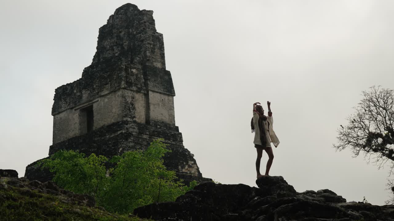 A woman stands on a rock with arms raised in front of a Mayan pyramid at Tikal, Guatemala, during overcast weather.
