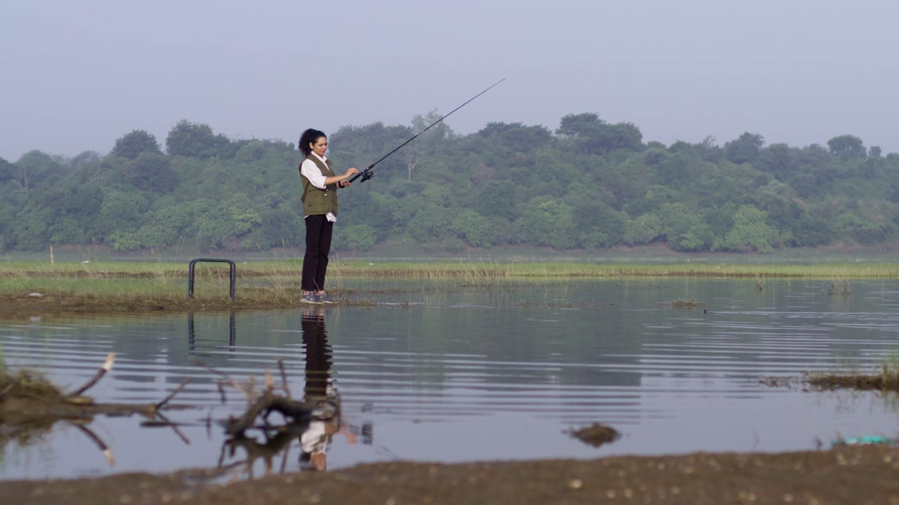 A South Asian girl wearing outdoor vest through shallow lake water holding her fishing rod after casting
