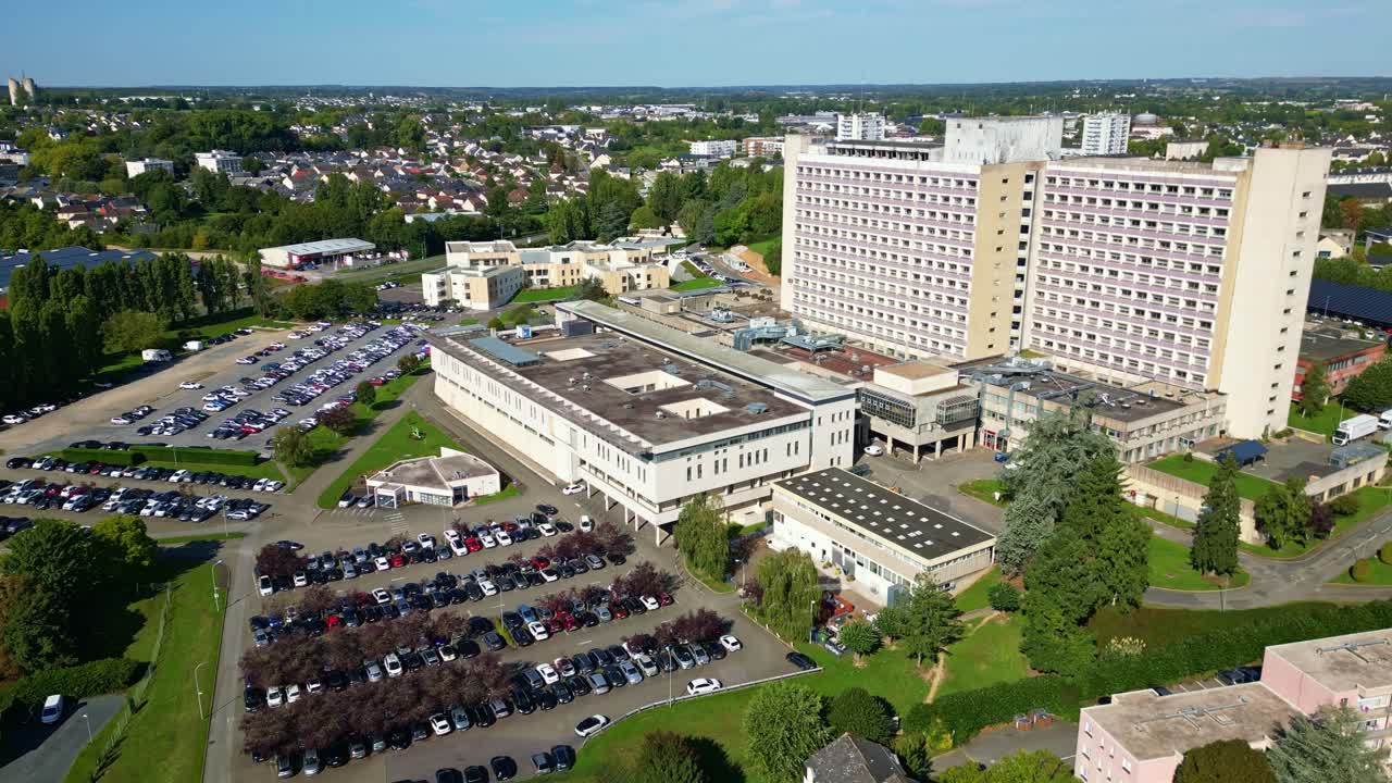 Drone shot over Laval showing the main hospital complex and its parking areas, with a wide view of the city in the background. The drone moves steadily, revealing the full layout of the buildings