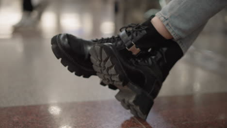leg view of poised woman seated on mall bench with crossed legs in chunky boots gently shaking, denim jeans visible, tiles and glass railing background, neutral lighting capturing gesture