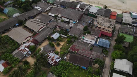 Drone-Captura-La-Vista-Aérea-De-Muchas-Casas-En-La-Ciudad-Cerca-De-La-Playa-De-Malibu-En-Koh-Phangan,-Tailandia,-Junto-Con-Un-Bote-En-El-Agua,-Una-Playa-De-Arena,-Corales-Y-Montañas