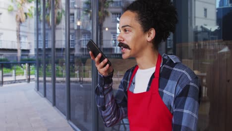barista de raza mixta con bigote que lleva un delantal hablando en un teléfono inteligente