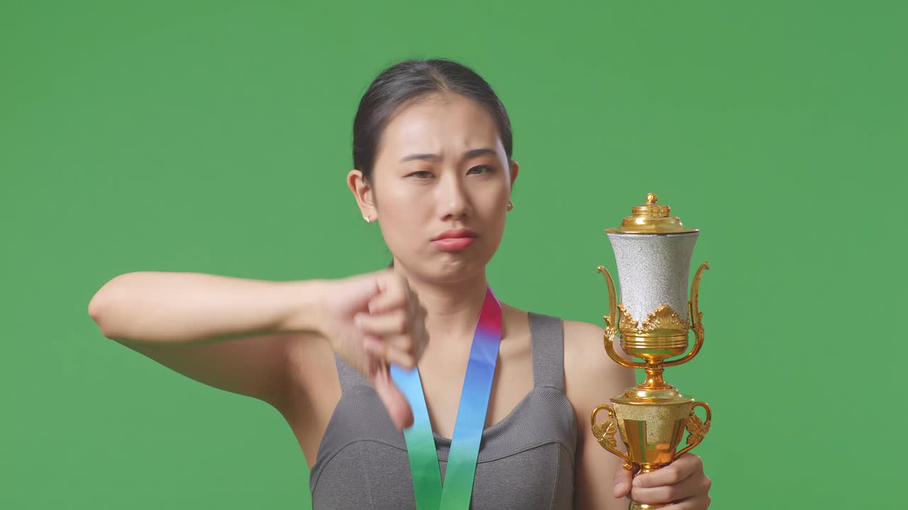 금메달과 트로피를 들고 있는 아시아 여성이 손가락을 아래로 가리키며 패배자를 조롱하는 몸짓을 하고 있다.
