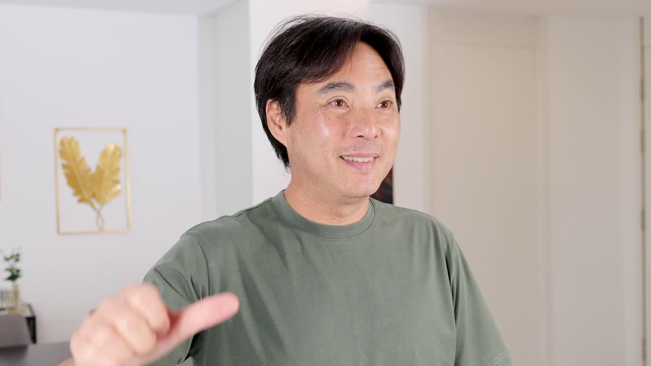 Asian man in green shirt indoors giving thumbs down, expressing disapproval in a bright room